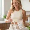 Woman in a cream athletic top scooping Perfect Collagen® Professional Grade powder into a glass of water in a bright, modern kitchen.